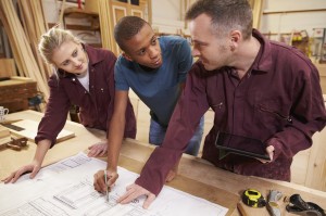 Carpenter With Apprentices Looking At Plans In Workshop